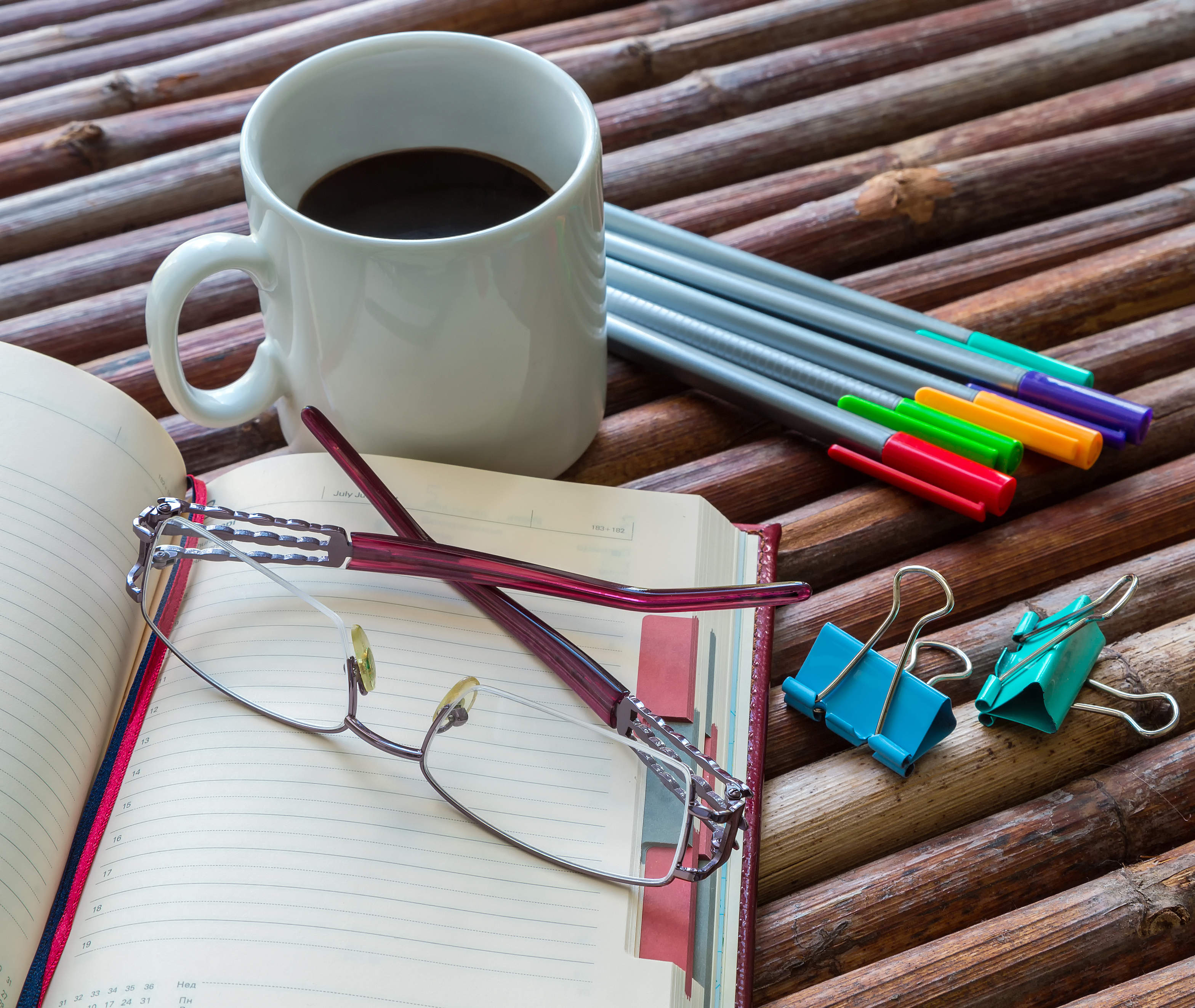 An open notebook, glasses, coffee in a mug, pens and clippers are laid out on a wooden surface